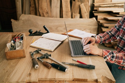 Person in plaid shirt sitting at table with laptop, notes, and tools.